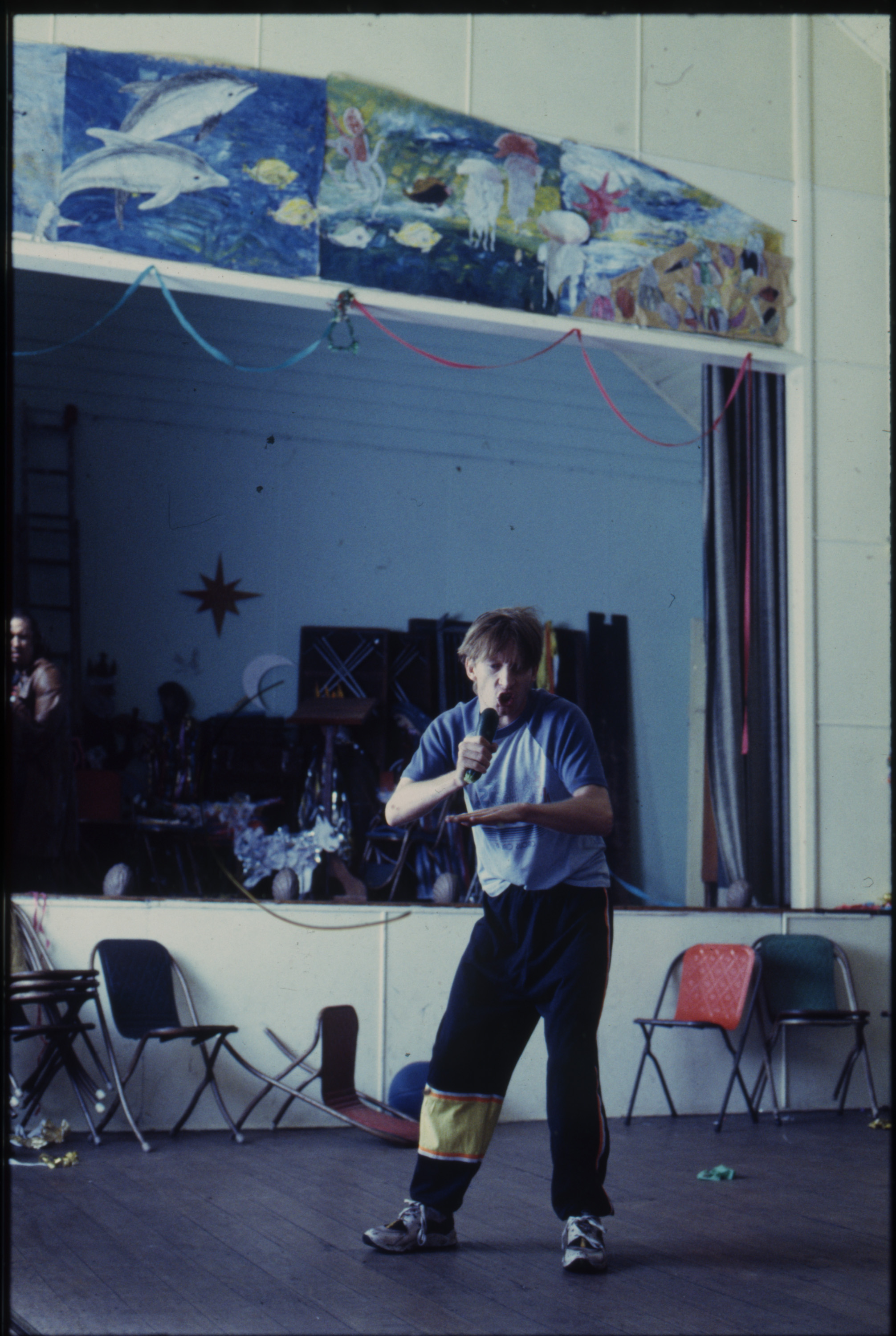 A man in his early 30s, wearing 90s-style sportswear, is in a community hall singing into a zucchini