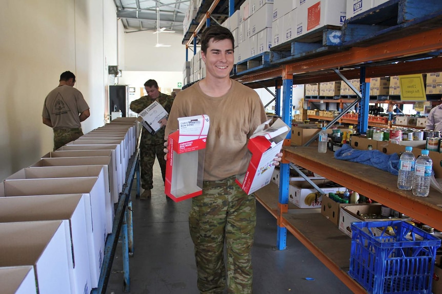 Men in army shirts carry boxes and pack food items into boxes in a warehouse.