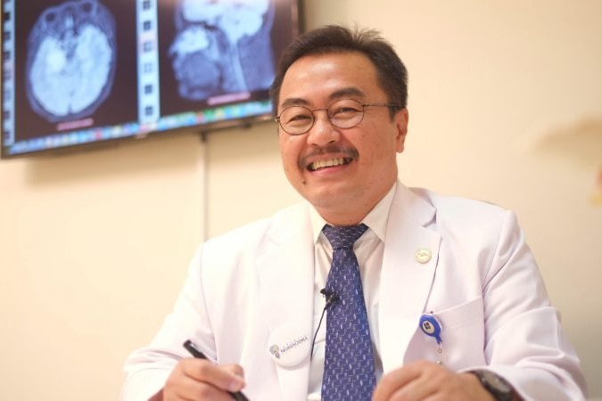 A bespectacled doctor in white lab coat smiles in front of images of brain scans