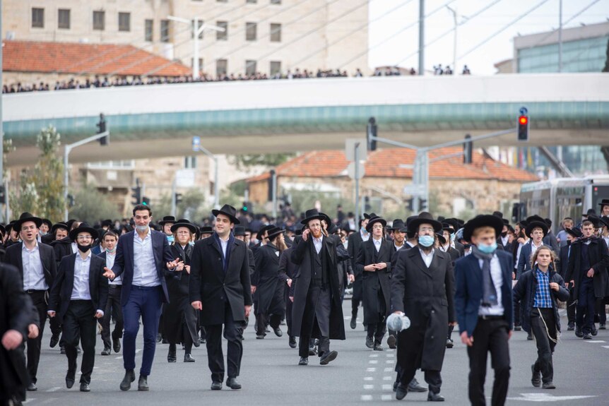 A large group of men in dark suits and hats walk en masse on a city street on a cloudy day.