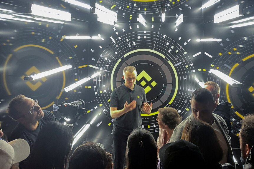 A man with short hair and glasses in a black polo shirt speaks in front of a hi-tech black backdrop as people record him.