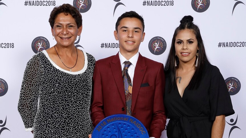 Jack Peris at the 2018 Naidoc awards, holding his award, standing between two women