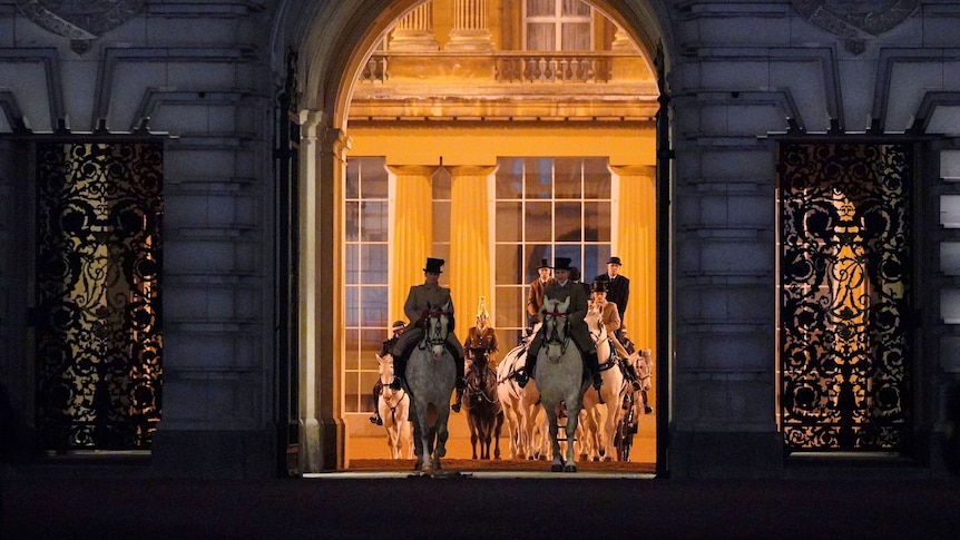 People ride on horseback through an archway out of the palace. The inside of the palace is lit, while outside is dark