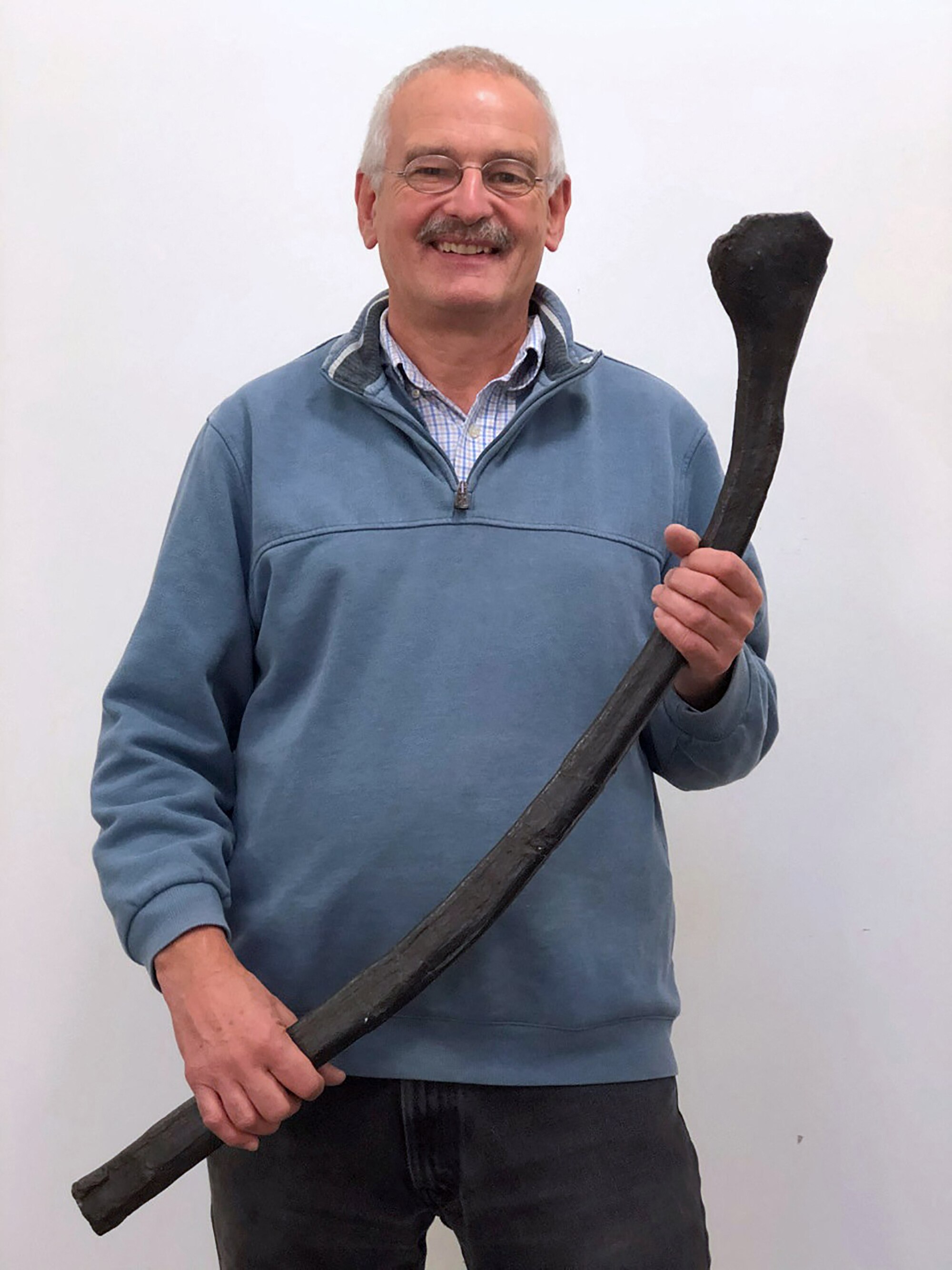 A grinning moustached man holds a large black long item that looks slightly like an oversized burn-out matchstick