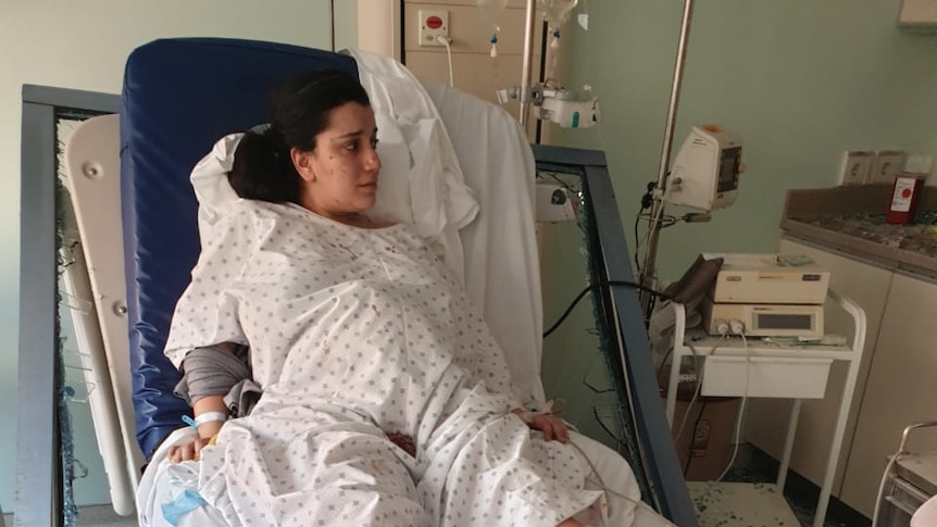 A woman with dark hair and wearing a hospital gown lays on a bed with shattered glass above her.