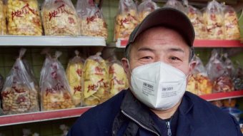 A man poses for a photo in a surgical mask in front of his snacks for sale.