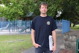 Darby Garrett stands in a park holding a newspaper looking at the camera with a children's playground in the background.