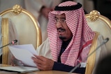 King Salman sits in a gold chair, reading from paper into a microphone