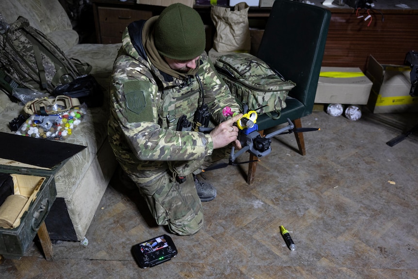 A soldier crouches over a drone, attaching brightly coloured tubes to the top of it 