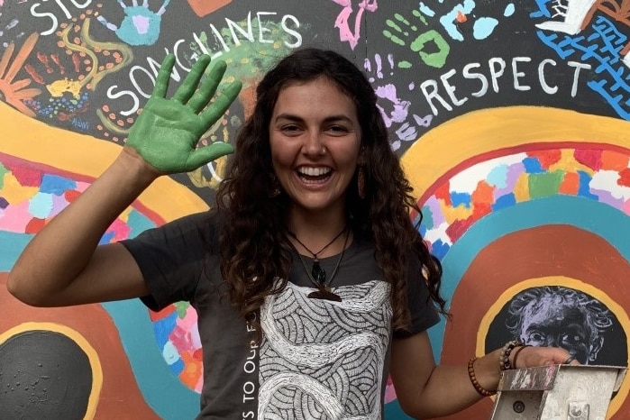 A young indigenous woman stands in front of a mural