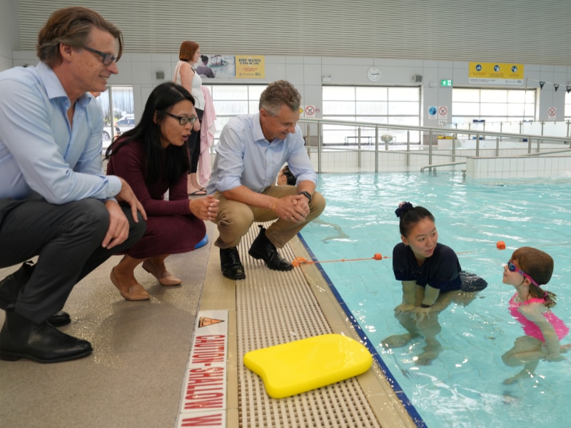 Personas vestidas de oficina se agachan junto a una piscina cubierta para hablar con un instructor de natación y un estudiante de natación