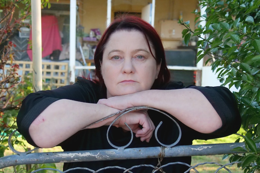 Sharon Henderson leans on her front gate, with her house behind, looking at the camera