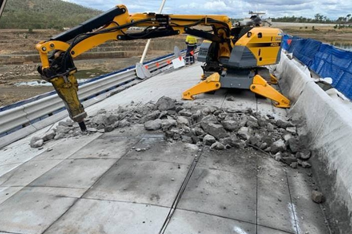 A yellow digger drills into the top of a thick concrete dam wall.