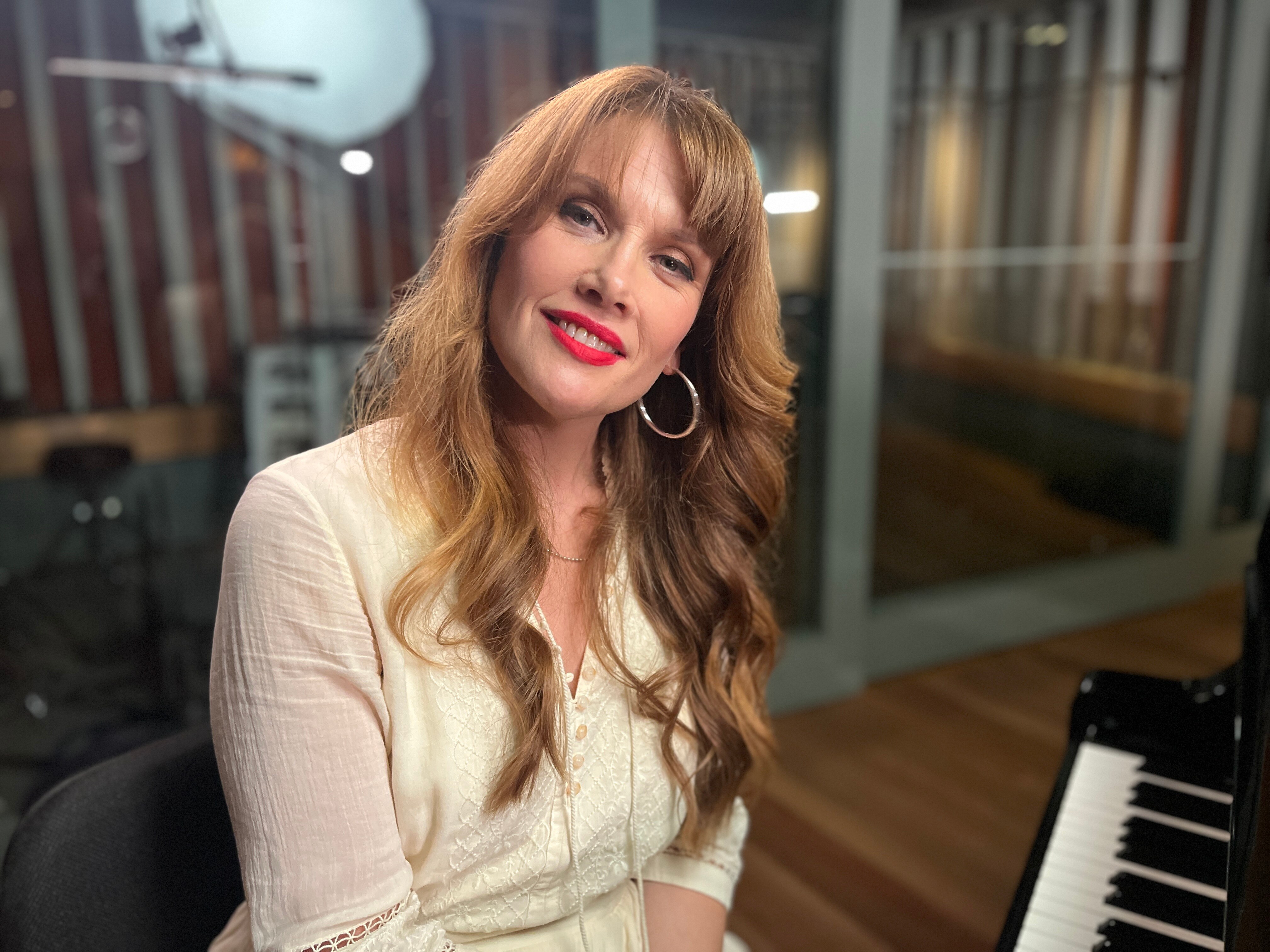 Woman in a white top and long wavy hair sits at a piano.