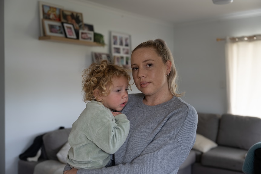 A woman holds her small child, looking into camera with a serious expression.