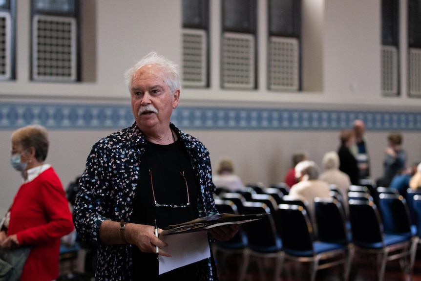 Older man with a clipboard in a hall filled with seats.