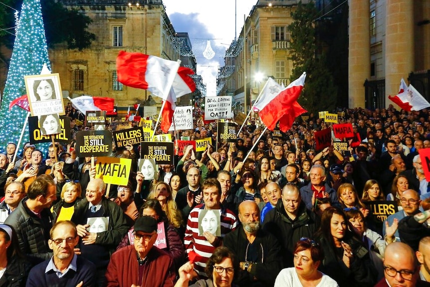 You look down at a large crowd of people waving the Maltese flag and holding signs that read 'Mafia' in a neoclassical square.