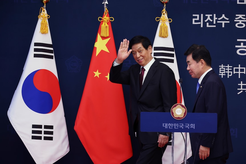 A clothed man waves to the camera as he and another man walk past the Chinese and South Korean flags.