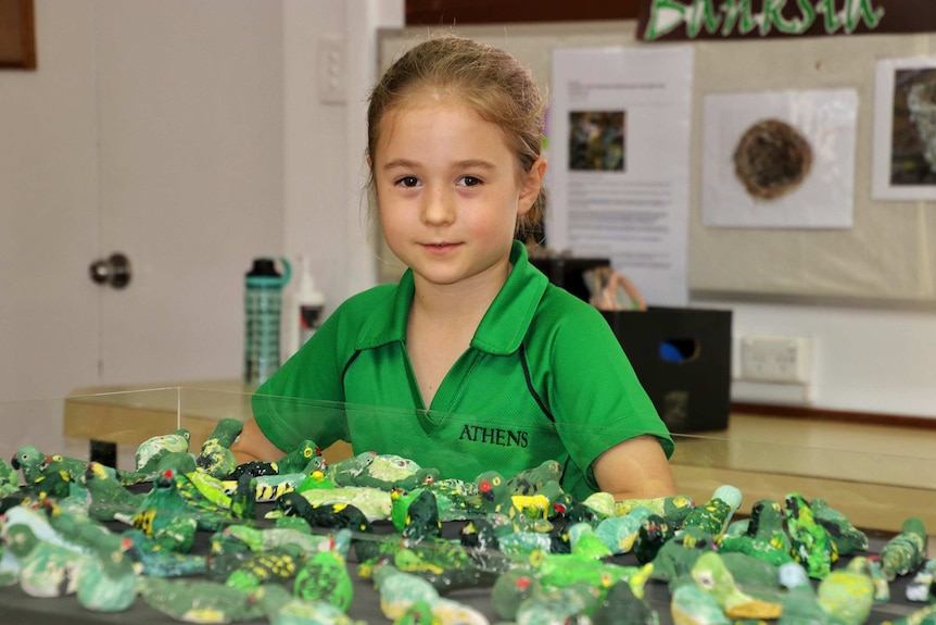 A girl in a green school uniform in front of a glass case with lots of small western ground parrot figures inside.