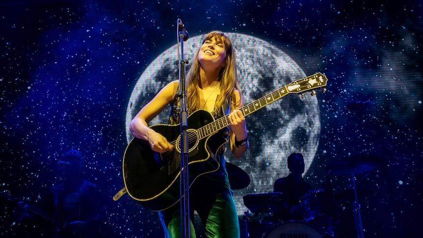 Missy Higgins stands in a green jumpsuit on stage holding a guitar, in front of a microphone with her eyes closed while singing.