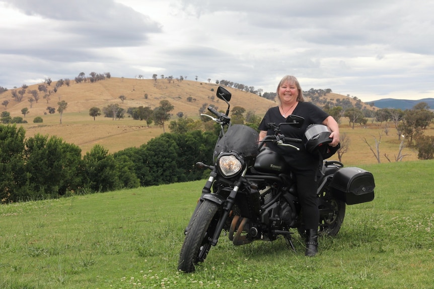 A middle-aged woman with blonde hair wearing all black clothing and sitting on a black motorcycle in the countryside.