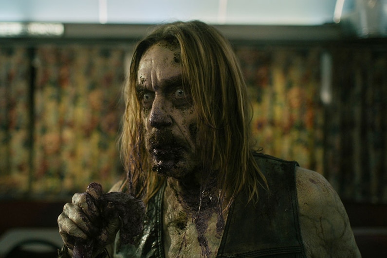 A zombie with blood splattered on face stands in diner with intense expression and holding an unknown object in hand.