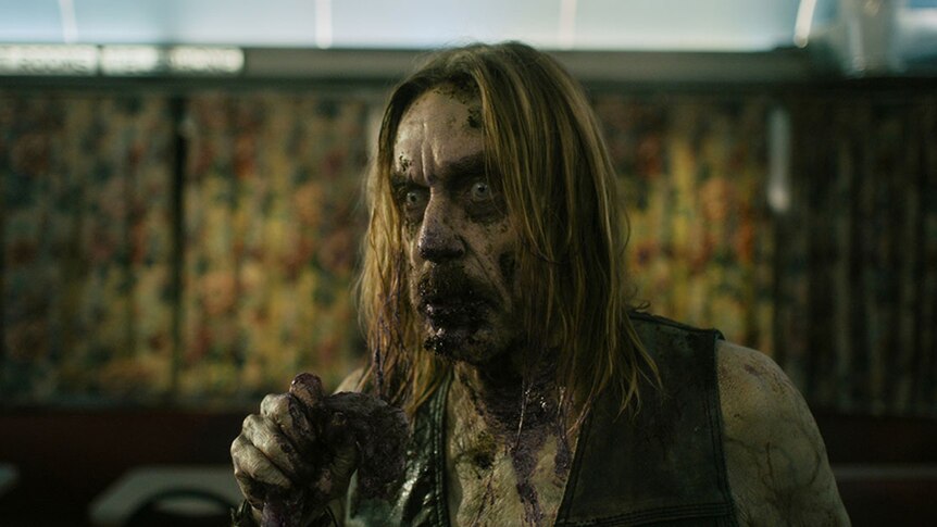 A zombie with blood splattered on face stands in diner with intense expression and holding an unknown object in hand.