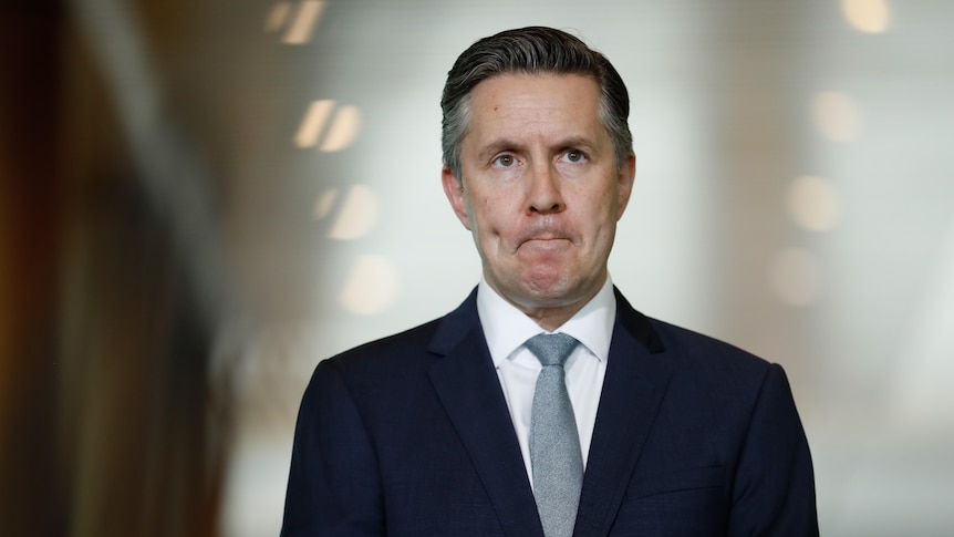 A man in a dark suit with a white shirt and grey tie frowns as he fronts media and the hallway behind him is blurred.