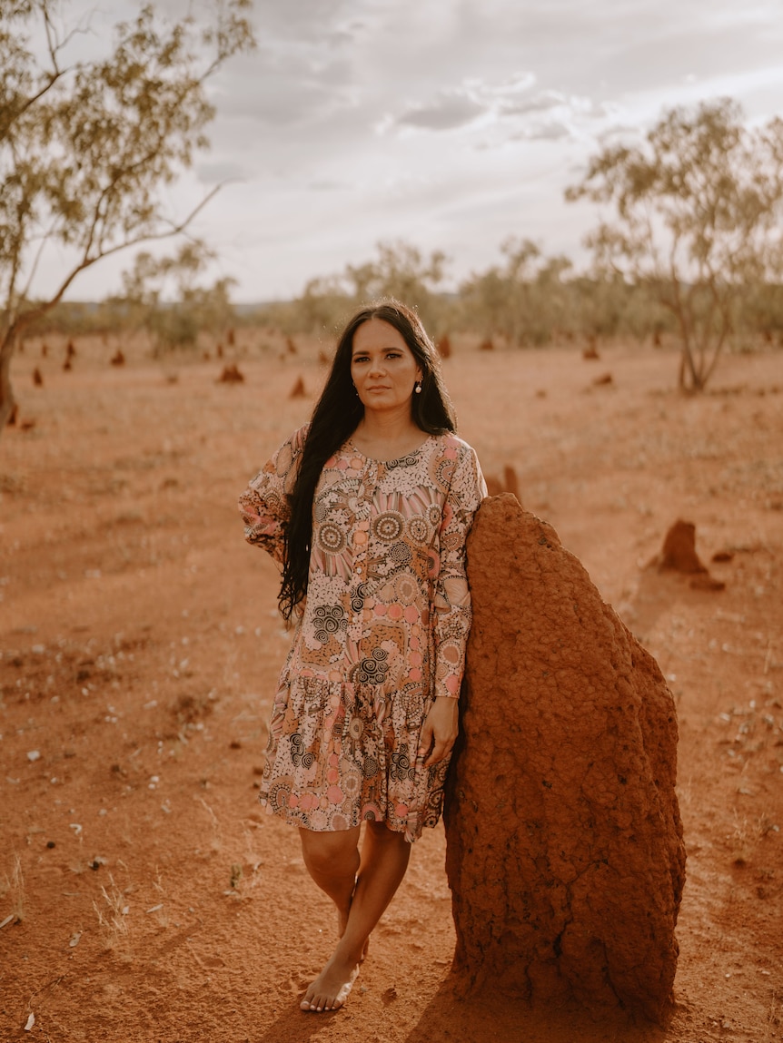 An Indigenous woman poses for a photo in a desert landscape wearing pink and neutral patterned dress