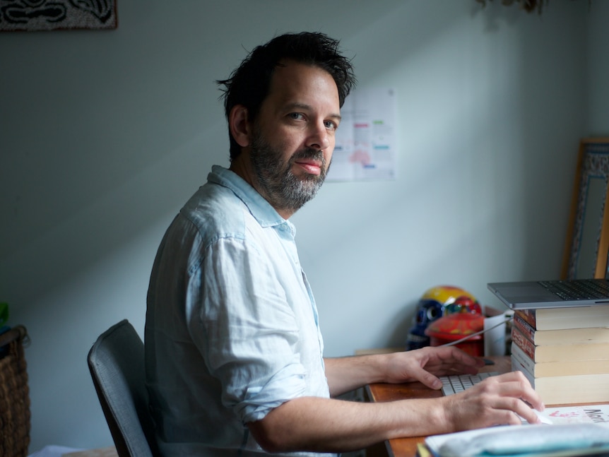 Antony Loewenstein sits at his desk by his computer and looks at the camera 