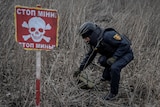 A red sign with a skull and cross bones stands in front of a man who is picking something up from a field.