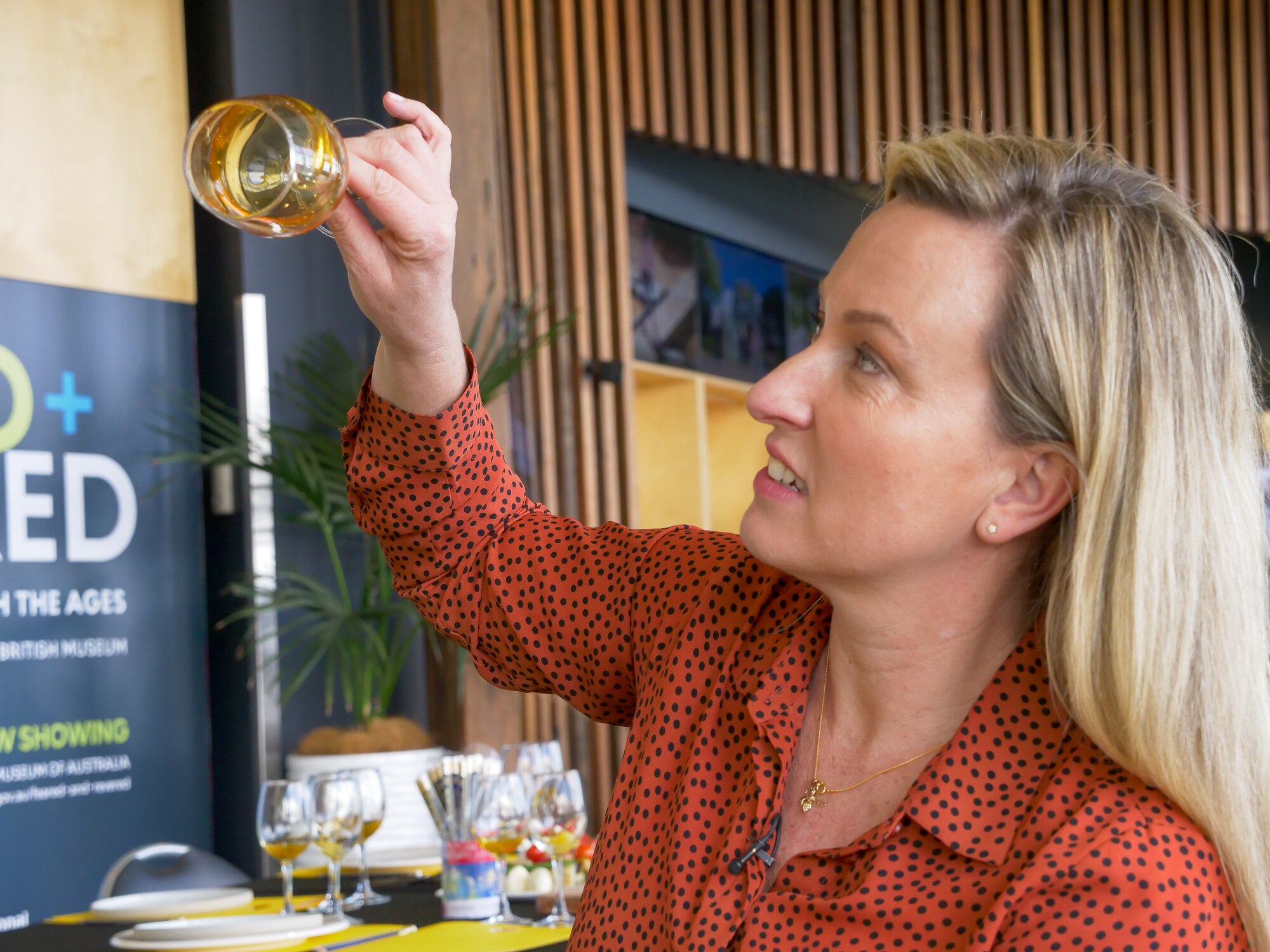 Jessica Locarnini, wearing a burnt orange top with black spots, studies honey inside a wine glass she is holding up.