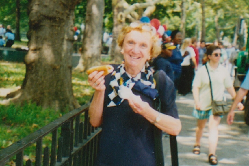 Yvonne Kennedy holds a hot-dog and smiles, standing on a busy footpath near a park.