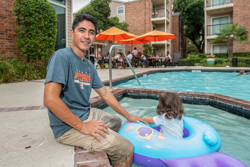 A man sits near a pool while his daughter plays in a floatie