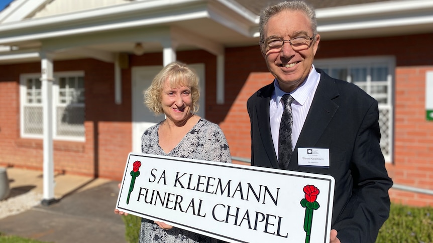 A smiling, well-dressed couple stands in front of a tidy red brick building hold a sign that reads SA Kleemann Funeral Chapel.