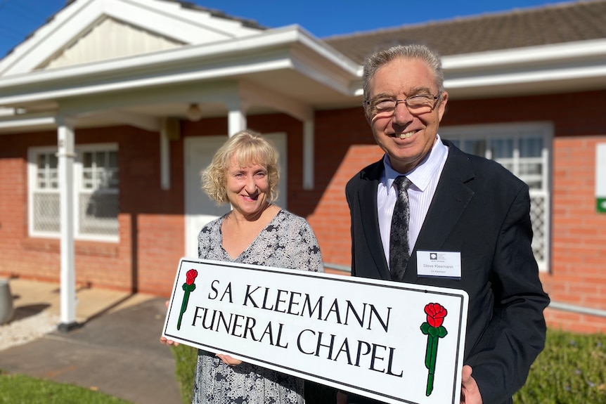 a woman and man standing in front of a red brick building, holding a sign reading S A Kleemann funeral chapel