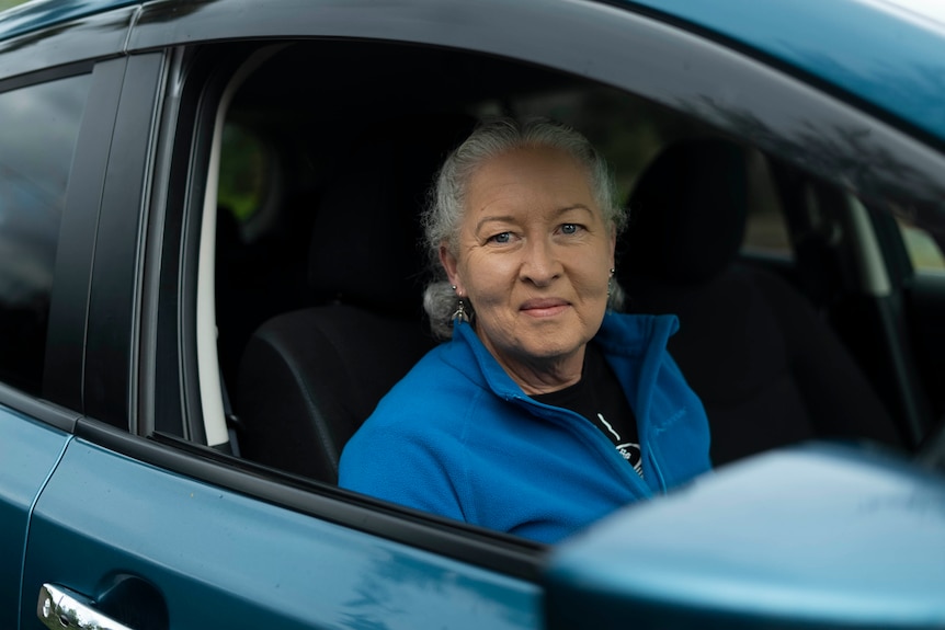 A woman with grey hair and a blue jumper sits behind the wheel of her electric car