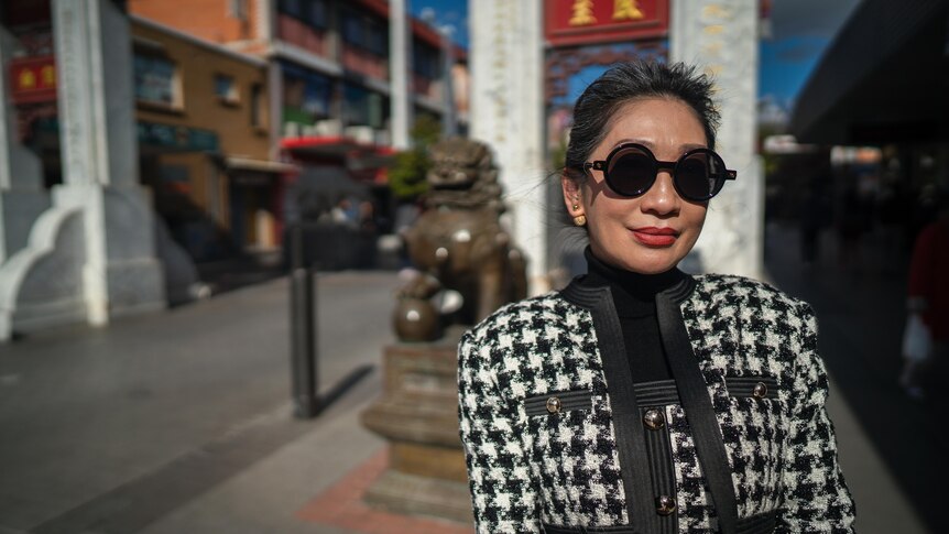 Asian woman wearing black and white houndstooth jacket with sunglasses standing in front of a bronze lion.