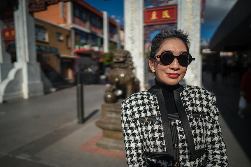 Asian woman wearing black and white houndstooth jacket with sunglasses standing in front of a bronze lion.