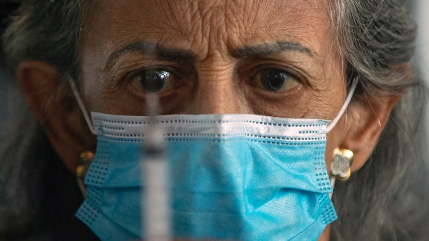 A older woman wearing a face mask eyes a syringe held in front of her face.
