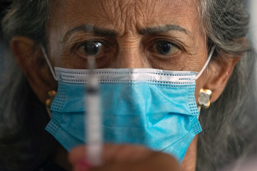 A older woman wearing a face mask eyes a syringe held in front of her face.
