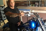 A middle-aged man with tattoos on his arms, wearing a black T-shirt and trousers, sitting on a blue motorcycle.