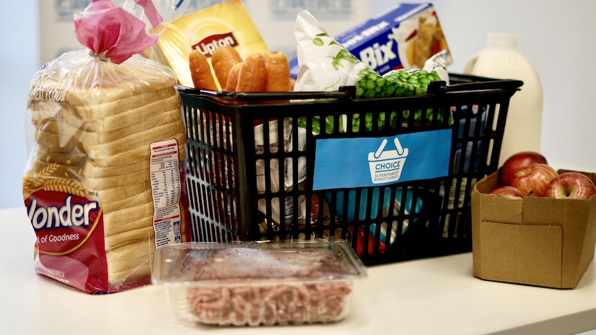 A black shopping basket sitting on a kitchen bench that contains bread, mince, carrots, apples and milk.