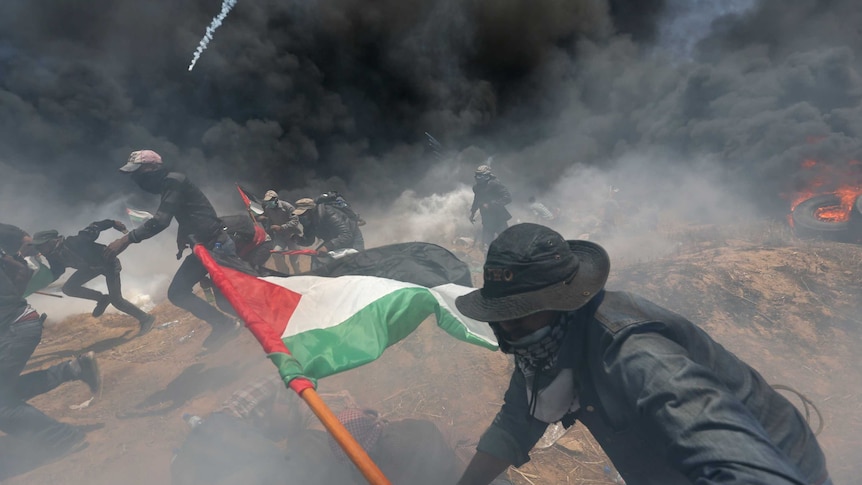 Palestinian demonstrators run for cover holding flags and with black smoke filling the air.