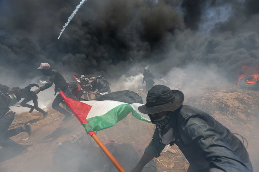 Palestinian demonstrators run for cover holding flags and with black smoke filling the air.