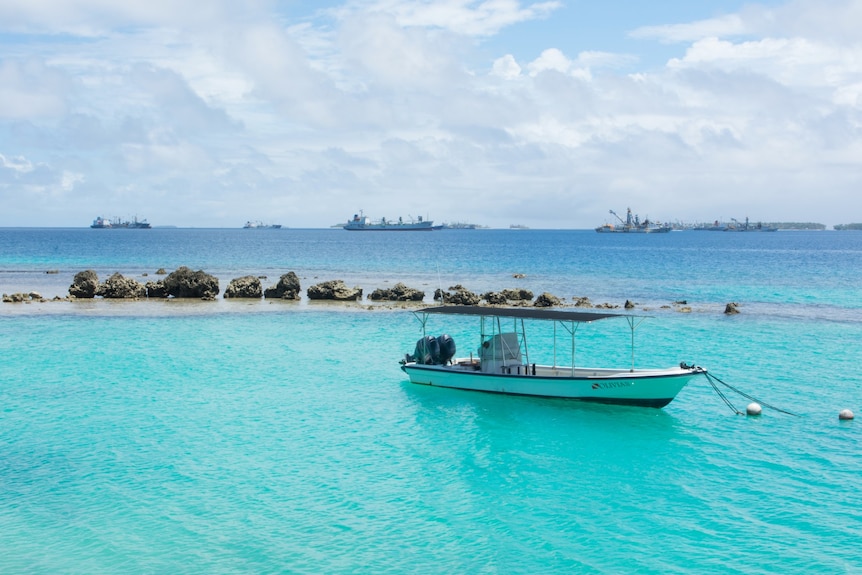 A small boat with an outboard motor on blue water, with large cargo ships in the distance