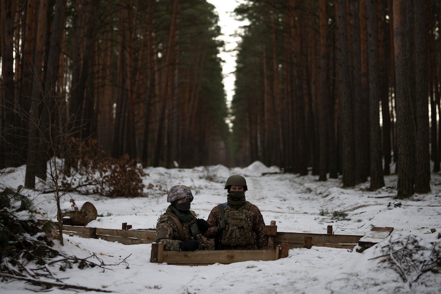 Two soldiers are half-way out of a hole in the snowy forest floor. 