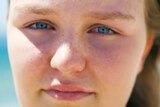 Brielle looks to the camera. Her blue eyes contrast with the blue of the sea behind her in a tight headshot.