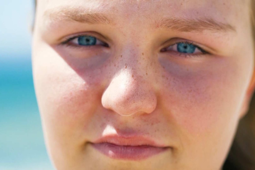 Brielle looks to the camera. Her blue eyes contrast with the blue of the sea behind her in a tight headshot.