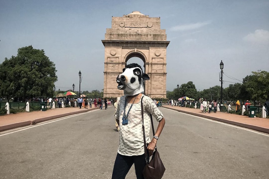A woman stands on a boulevard wearing a cow mask in front of New Delhi's India Gate.